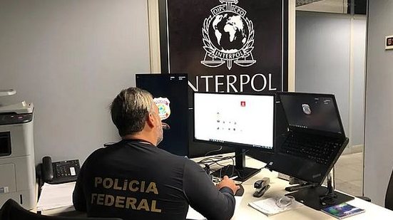 Imagem ilustrativa de um agente da Polícia Federal no escritório com a logo da Interpol | Polícia Federal/Divulgação