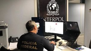 Imagem ilustrativa de um agente da Polícia Federal no escritório com a logo da Interpol | Polícia Federal/Divulgação